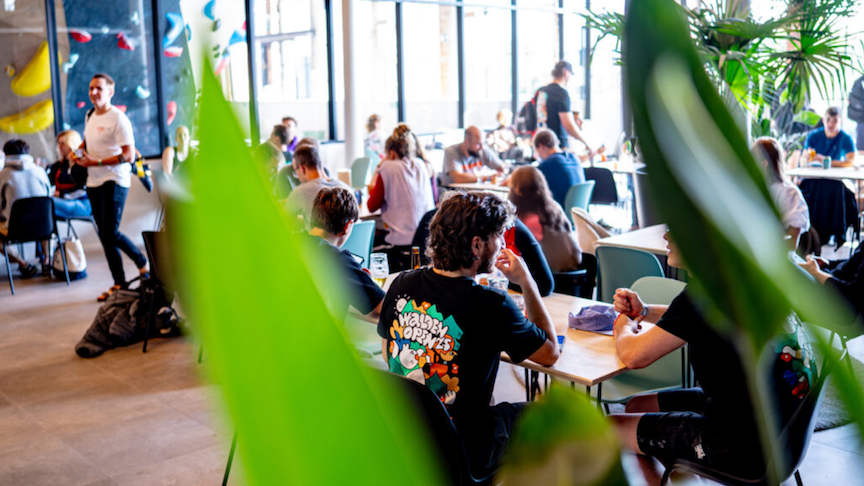 Klimzaal bar Walden Galveston Gent: Gezellige en drukke klimzaal bar van Walden, met klimmers die genieten van drankjes en gesprekken aan de tafels, gezien door een grote groene plant.