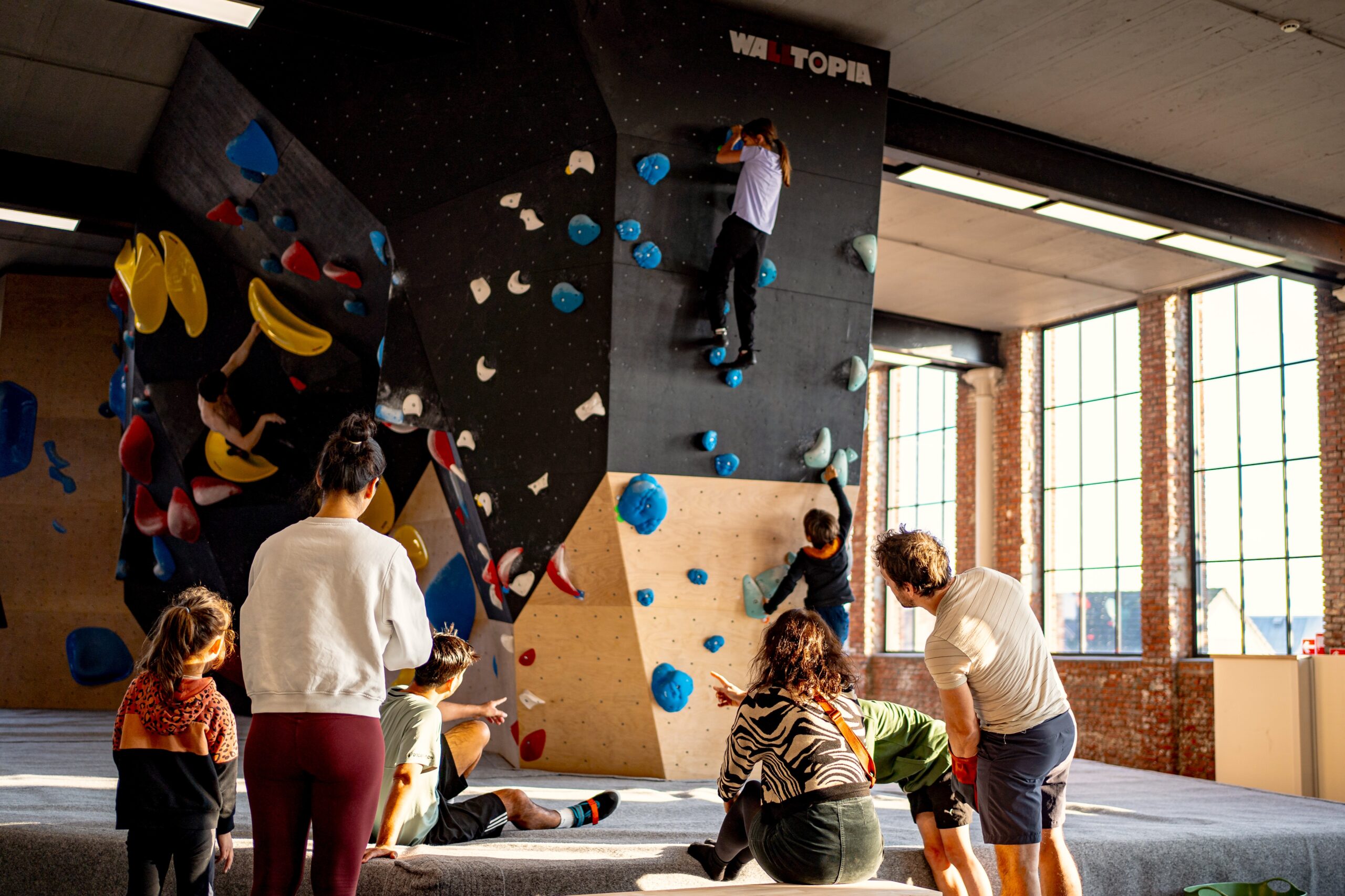 A lively group enjoys indoor bouldering at a modern climbing gym named 'Walltopia.' The spacious facility features diverse climbing walls equipped with colorful handholds and footholds, catering to climbers of all ages and skill levels. In the foreground, children and adults observe and discuss climbing techniques, while in the background, climbers actively scale the walls. The bright, natural light streaming through large windows creates an inviting and energetic atmosphere, perfect for fitness enthusiasts, families, and adventure seekers. Ideal for content related to indoor climbing, family activities, fitness, and recreational sports.